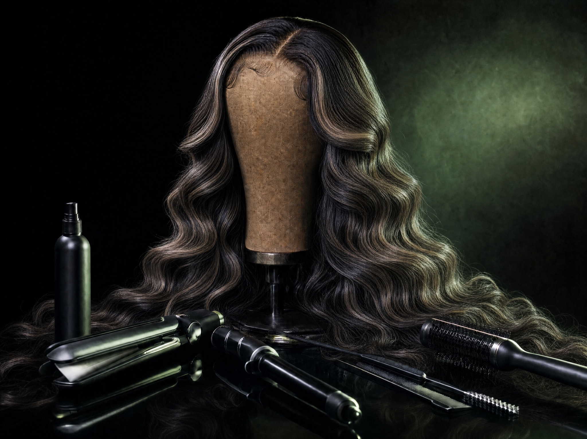 A long, glossy, highlighted lace-front wig styled in voluminous waves on a canvas block head, surrounded by styling tools in an atelier still life.